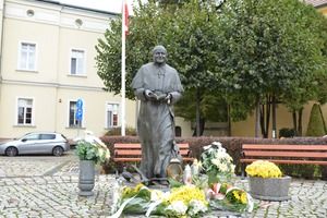 Pomnik Papieża Jana Pawła II na placu Niezłomnych w Kościanie.  Przed pomnikiem zapalone znicze oraz kwiaty, w tle drzewa oraz zabudowania. (photo)